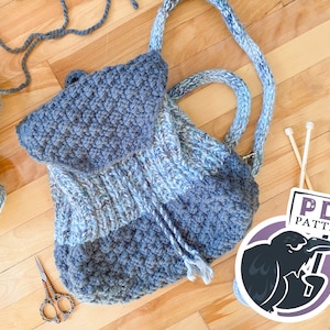 May include: A blue and white crocheted tote bag with a fringe detail. The bag is on a wooden surface with yarn and knitting needles. A sticker with the text "PDF PATTERN" and a crow illustration is in the lower right corner.