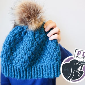 May include: A blue knitted beanie with a fluffy brown pom pom. The beanie is being held up in front of a person's face. A sticker with a crow and the text "PDF PATTERN" is in the lower right corner of the image.