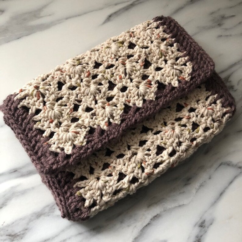 May include: A crocheted clutch bag with a brown border and a cream colored body with a floral pattern.