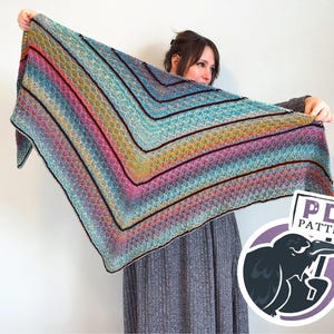 May include: A colorful, handmade shawl with a chevron pattern. The shawl features a gradient of colors, including pink, blue, yellow, and black. The shawl is held up, showcasing its size and design. A PDF pattern logo is in the bottom right corner.
