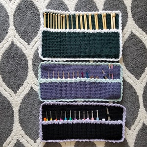 May include: Three crocheted needle holders with different colored fabric and white trim. The top holder is green, the middle is purple, and the bottom is black. Each holder has multiple slots for needles.