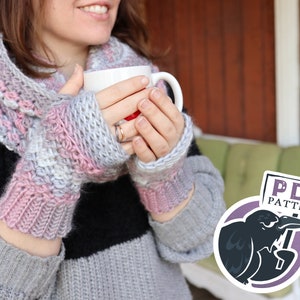 May include: A pair of pink and gray crocheted fingerless gloves with a white crocheted infinity scarf. The gloves and scarf are worn by a person holding a white mug. A sign with the text "PDF PATTERN" is held by a cartoon crow.