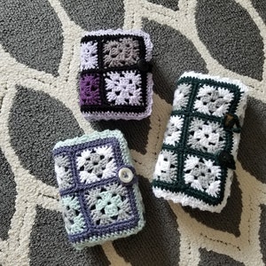 May include: Three crocheted square-shaped pouches in different color combinations. The pouches are made with white, black, gray, purple, and light green yarn. The pouches are on a gray and white patterned rug.
