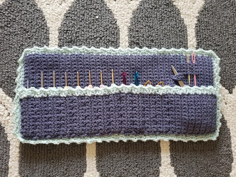 May include: A crocheted needle holder with a light blue border and a dark blue center. The holder has multiple slots for needles and other tools.