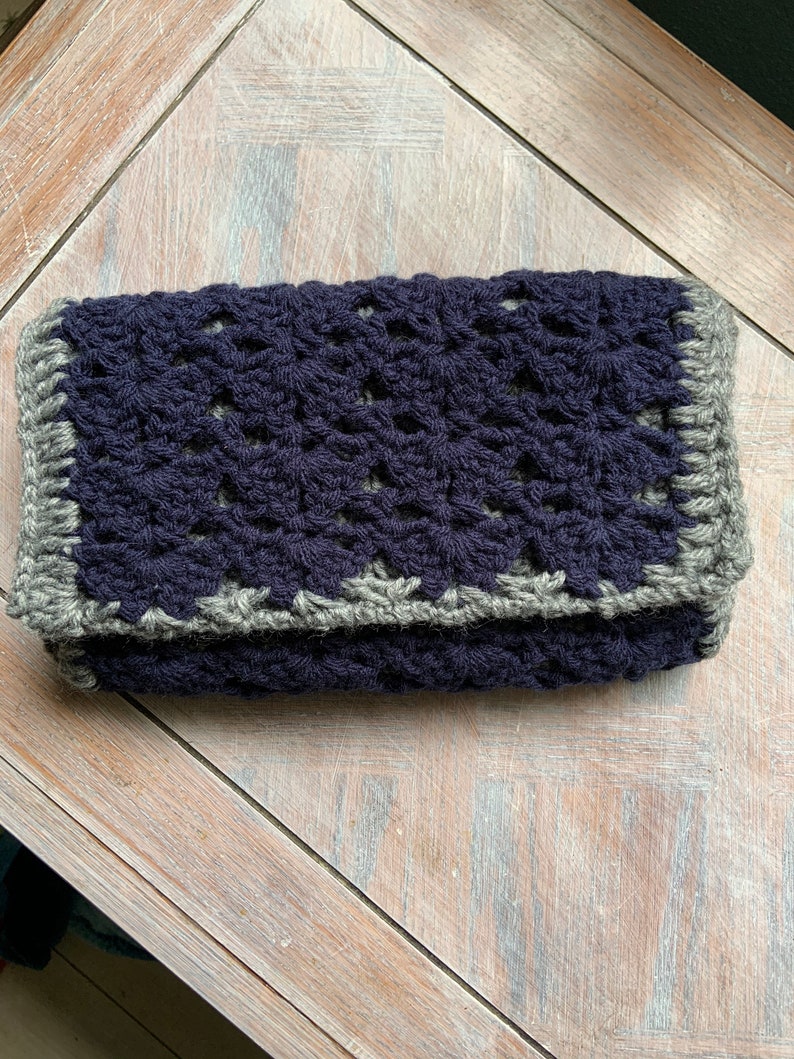 May include: A crocheted clutch bag with a dark blue body and a gray border.
