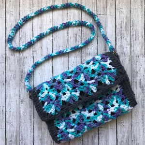 May include: A crocheted handbag with a gray, white, blue, and purple color pattern. The bag has a long strap and a flap closure.