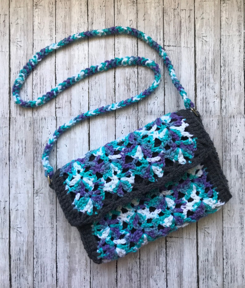 May include: A crocheted handbag with a gray, white, blue, and purple color pattern. The bag has a long strap and a flap closure.