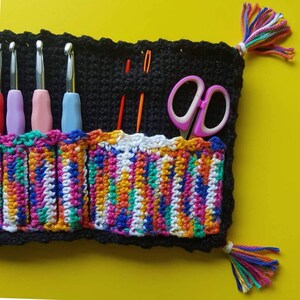 May include: A colorful crocheted knitting needle holder with a black crocheted base. The holder has six slots for needles and a pocket for scissors. The holder has a fringe on each side.