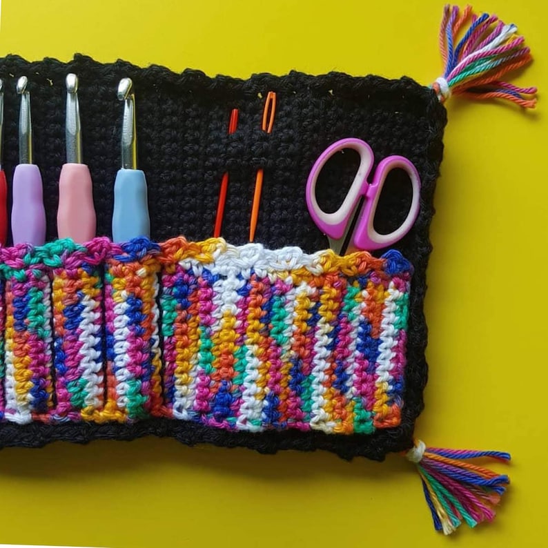 May include: A colorful crocheted knitting needle holder with a black crocheted base. The holder has six slots for needles and a pocket for scissors. The holder has a fringe on each side.