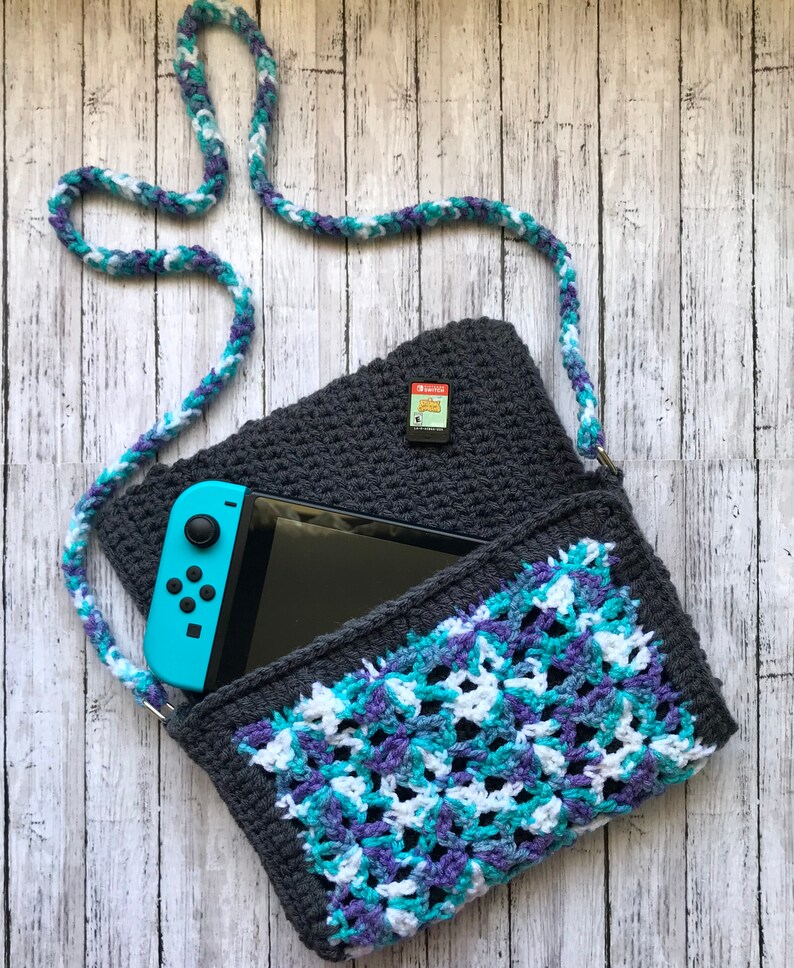 May include: A crocheted gray and blue bag with a white strap. The bag has a pocket on the inside that can hold a Nintendo Switch console. A game cartridge is visible on the top of the bag.