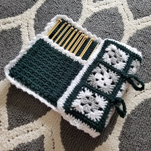 May include: A crocheted needle case with a green and white design. The case has a pocket for needles and a flap that closes with ties. The case is lying on a gray and white patterned rug.