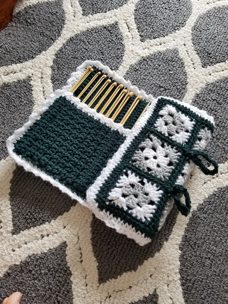 May include: A crocheted needle case with a green and white design. The case has a pocket for needles and a flap that closes with ties. The case is lying on a gray and white patterned rug.
