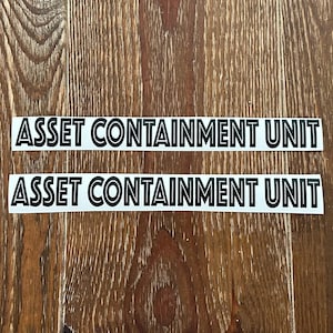 May include: Two white stickers with black text that reads "ASSET CONTAINMENT UNIT" are displayed on a wooden surface. The stickers are rectangular and have a clean, modern font. The wood grain background is a warm brown color.