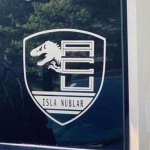 May include: A white vinyl sticker with a black shield design. The shield features a dinosaur silhouette and the text "ISLA NUBLAR".