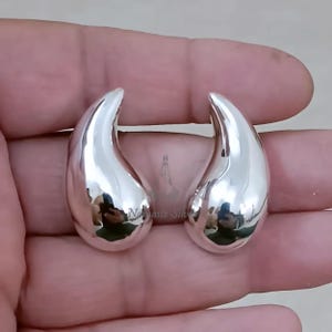 Può includere: Un paio di orecchini a goccia in argento lucido. Gli orecchini hanno una superficie liscia e riflettente e una forma curva e allungata. Sono esposti su uno sfondo neutro.
