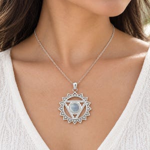 May include: A silver pendant necklace featuring a circular design with a central round blue stone set within a triangular frame, surrounded by a petal-like border. The pendant hangs from a delicate silver chain against a white textured fabric.