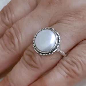 May include: A silver ring with a large, round, white pearl. The pearl is set within a textured, rope-style border. The ring's band has a twisted design. The ring is shown on a person's finger.
