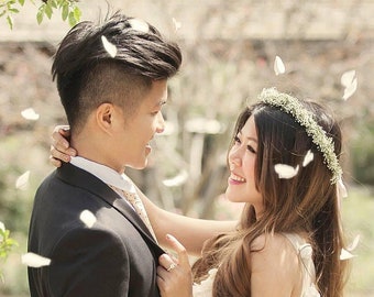 May include: A couple in wedding attire, the groom in a black suit and the bride in a white dress, are embracing and looking at each other. White flower petals are falling around them.