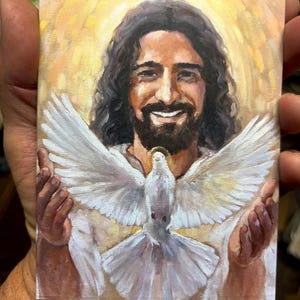May include: A small rectangular painting depicts a smiling figure with long dark hair and a beard, surrounded by a golden halo. A white dove with outstretched wings is positioned in front of the figure. The artwork is held by a person.