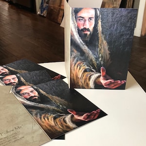 May include: Greeting cards featuring a portrait of a man with a beard, reaching out his hand. The artwork is in a painterly style with warm tones and a dark background. The cards are displayed in a stack and upright.