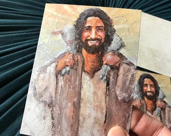 May include: A painting of a man with long brown hair and a beard, wearing a brown robe, carrying a white lamb on his shoulders. The background is a light brown with a sun shining in the distance.