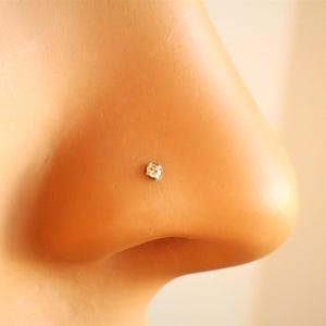 Peut inclure: Gros plan sur un nez avec un petit clou de nez en forme de carré, en pierre précieuse transparente. Le clou est serti dans un métal de couleur claire et est positionné sur la narine droite. Le teint est clair.