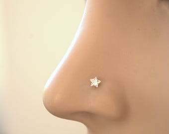 May include: A close-up of a nose with a small, star-shaped nose stud. The stud is silver-colored and has a five-pointed star design. The skin tone is light, and the background is blurred.