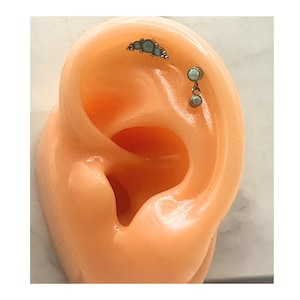 Puede incluir: Modelo de oreja con dos joyas. Una es una barra curva con pequeñas piedras azul claro. La otra es un pendiente colgante con dos piedras azul claro. La joyería es de color plateado.