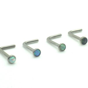 Peut inclure: Une collection de six piercings de nez en forme de L, ornés de petites pierres d'opale rondes et colorées. Les clous sont en métal argenté et sont disposés en rangée sur fond blanc. Les opales sont de couleurs variées.