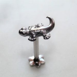 Puede incluir: Joyería corporal de plata con un diseño de lagarto detallado. El lagarto está mirando hacia la izquierda con la cola enroscada. La joyería es de estilo barra con una base en forma de flor.