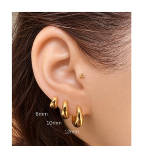 May include: Three gold hoop earrings in graduated sizes of 6mm, 10mm, and 12mm, along with a small gold stud earring featuring a three-dot cluster design. The earrings have a smooth, polished finish and are arranged in a row on the earlobe.