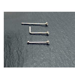 K&ouml;nnte beinhalten: Drei silberfarbene Nasenstecker, die horizontal angeordnet sind. Der obere Stecker ist gerade, der mittlere hat eine L-f&ouml;rmige Biegung und der untere Stecker endet in einer kleinen Kugel. Jedes St&uuml;ck ist mit einem klaren, runden Stein verziert.