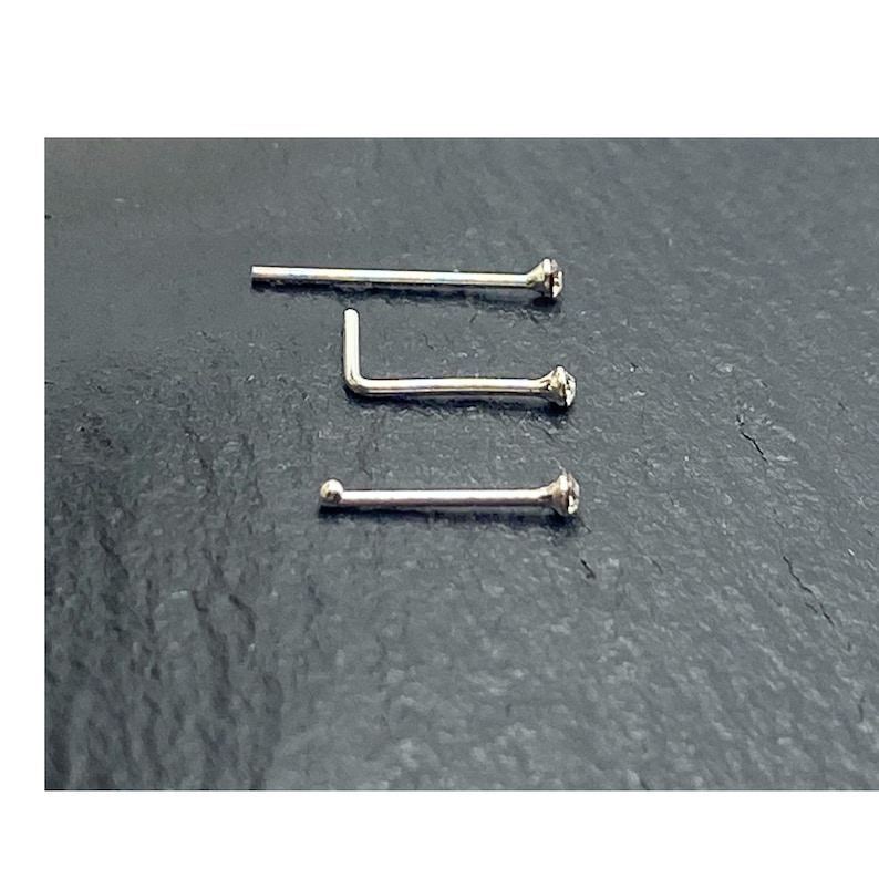 K&ouml;nnte beinhalten: Drei silberfarbene Nasenstecker, die horizontal angeordnet sind. Der obere Stecker ist gerade, der mittlere hat eine L-f&ouml;rmige Biegung und der untere Stecker endet in einer kleinen Kugel. Jedes St&uuml;ck ist mit einem klaren, runden Stein verziert.
