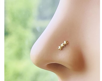 May include: A close-up of a nose with a gold nose stud featuring three small star-shaped charms. The nose stud is a bright gold color and is placed on the side of the nose.