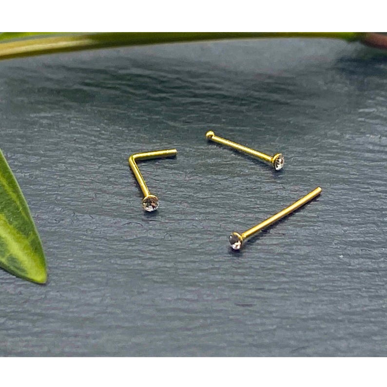 K&ouml;nnte beinhalten: Drei goldfarbene Nasenstecker, die jeweils mit einem kleinen, klaren, runden Schmuckstein am Ende versehen sind. Ein Stecker ist L-f&ouml;rmig, w&auml;hrend die anderen beiden gerade Stifte sind. Das Metall hat eine polierte Oberfl&auml;che und die Steine sitzen fest.