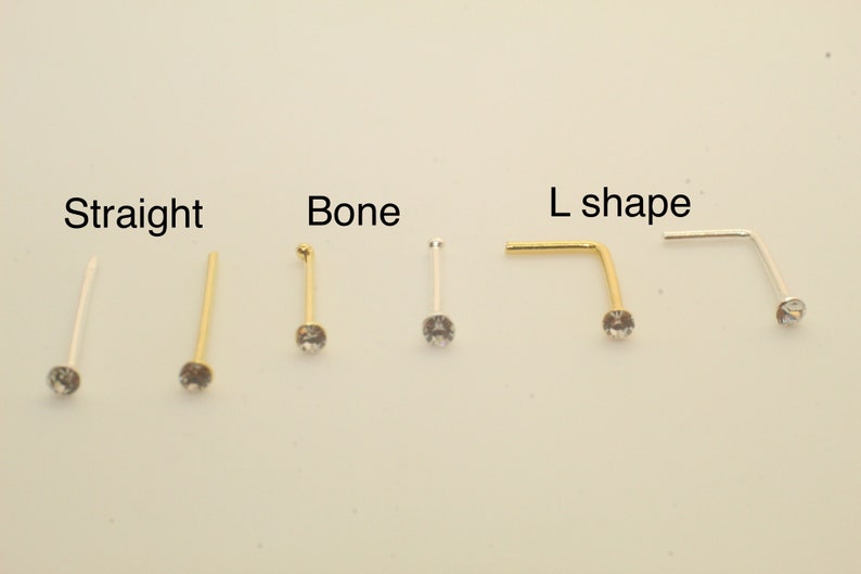 K&ouml;nnte beinhalten: Sechs Nasenstecker mit klaren Kristallen, angeordnet in Paaren mit den Beschriftungen Straight, Bone und L shape. Die Stecker haben gold- und silberfarbene Metallstifte in drei verschiedenen Formen, jeweils mit einem kleinen, runden Kristall.