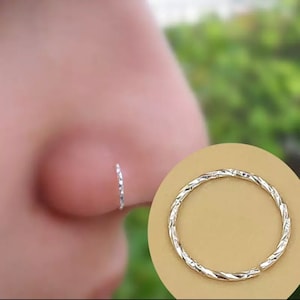 May include: A silver nose ring with a twisted design. The ring is shown on a model's nose and also on a white background.