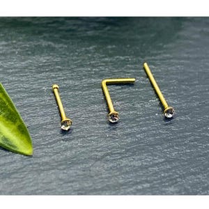 K&ouml;nnte beinhalten: Drei goldfarbene Nasenstecker, die jeweils mit einem kleinen, klaren, runden Schmuckstein am Ende versehen sind. Ein Stecker ist gerade, einer ist L-f&ouml;rmig und einer ist ein langer, gerader Stift. Das Metall hat ein poliertes Finish und die Steine.