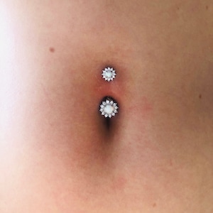 May include: A silver belly button ring with two clear flower-shaped charms. The charms have a white centre and are surrounded by small clear stones.