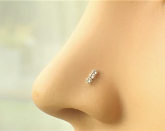 May include: A silver-toned nose stud featuring three small, clear, round-cut stones arranged in a vertical line. The jewelry is positioned on the side of a nose, showcasing the metallic setting and the reflective quality of the three stones.