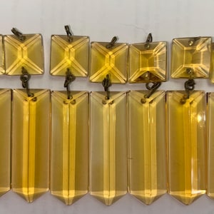May include: A collection of vintage amber-colored crystal chandelier prisms. The prisms are rectangular and square-shaped, with pointed bottoms and decorative metal connectors. The crystals are arranged in two rows, showcasing their reflective qualities.