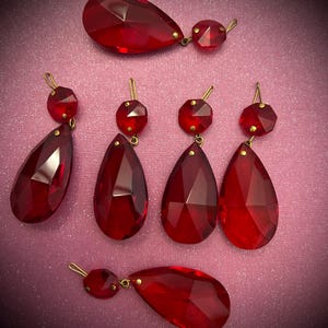 May include: A selection of faceted, ruby-red crystal pendants and earrings. The earrings have a teardrop shape with a smaller, round accent. The pendants and earrings have gold-coloured metal findings. The pieces are arranged on a pink background.