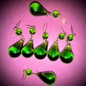 May include: A collection of green glass teardrop earrings with gold-colored hooks and accents. The earrings are arranged on a pink background, showcasing their vibrant color and unique design. The earrings have a shiny, transparent appearance.