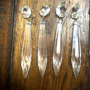 May include: Four clear glass chandelier prisms, each featuring a faceted octagonal top piece connected by a small gold-toned metal ring to a long, tapered, diamond-cut pendant. The glass components are transparent and reflect light through their facets.