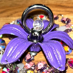 Op de afbeelding: Een paarse bloemvormige ringhouder met een zilveren ring en een heldere kristallen kraal. De ringhouder is omgeven door een kralenketting met iriserende en paarse kralen. De achtergrond is een houten oppervlak.