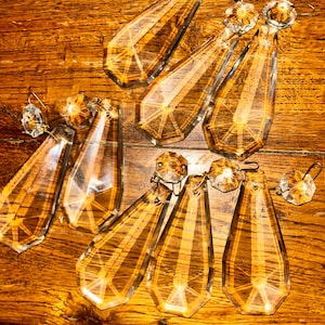 Puede incluir: Una colección de prismas de araña de cristal transparente con detalles metálicos dorados. Los prismas, en forma de lágrima, están facetados, reflejando la luz. Se disponen sobre una superficie de madera, mostrando su intrincado diseño y brillo.