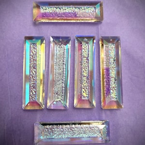 May include: Six rectangular glass pendants featuring a clear, beveled edge and a central textured, iridescent strip that shifts between purple, blue, and silver tones. Each piece has a small hole at the top for hanging, arranged in a grid pattern.