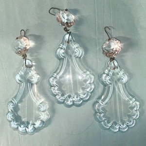 May include: Three clear glass chandelier crystals with a teardrop shape and scalloped edges. Each crystal is topped with a faceted, pink-tinted glass bead and a metal hook for hanging. The crystals are suspended against a light blue background.