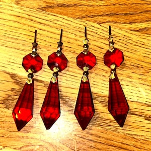 Puede incluir: Cuatro colgantes de araña de cristal rojo con detalles dorados. Cada colgante tiene una parte superior octogonal facetada, un conector dorado y una parte inferior en forma de diamante. Los colgantes están dispuestos sobre una superficie de madera.