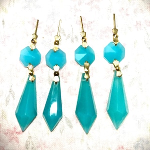 May include: Four pairs of turquoise-colored earrings. Each earring features a faceted, light blue gemstone pendant with a gold-colored metal accent. The earrings have a long, teardrop shape and are displayed against a floral patterned background.